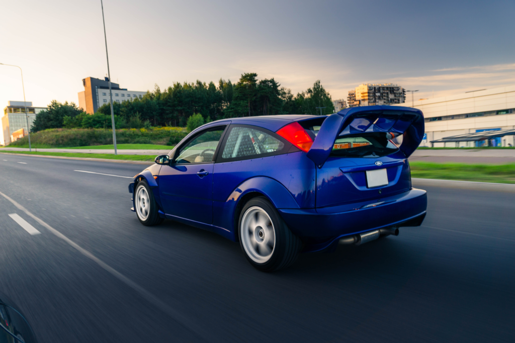 Mėlynas modifikuotas Ford Focus hečbekas su dideliu spoileriu važiuojantis gatve, iliustruojantis sportinių automobilių supirkimą Kaune su AutoDylu.