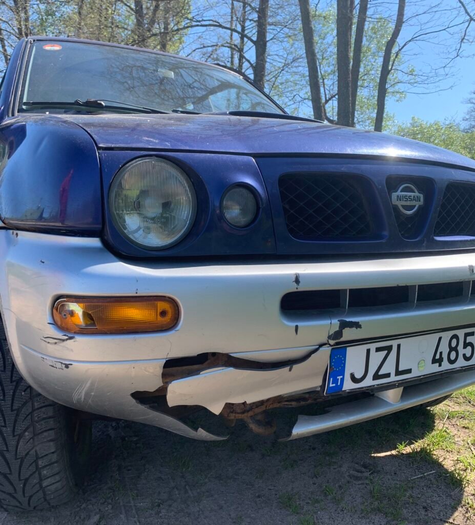 Mėlynas Nissan visureigis su sudaužytu priekiniu bamperiu po virtimo ant šono.